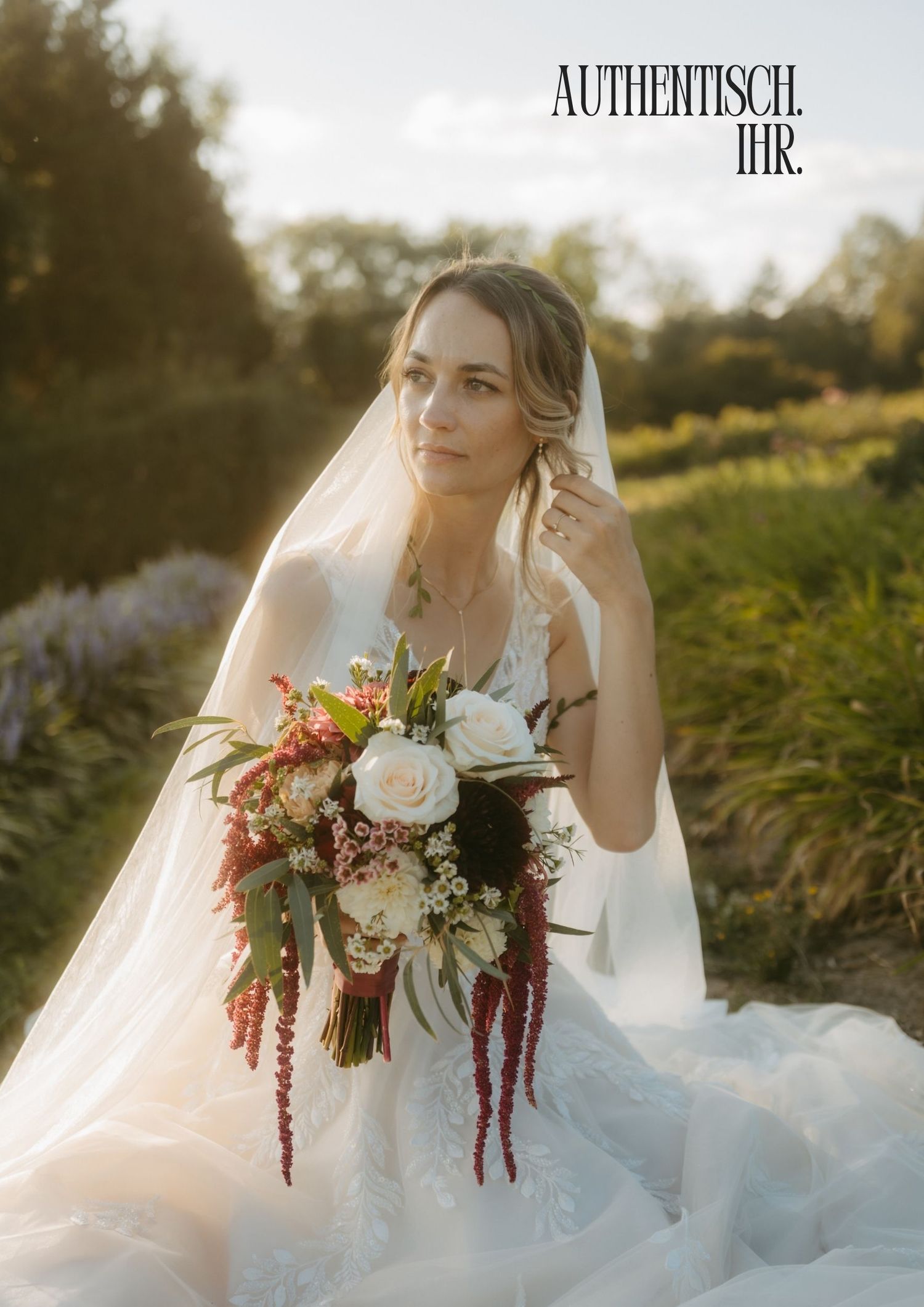 A bride in a white gown and veil holds a bouquet of white and red flowers outdoors in golden hour light.