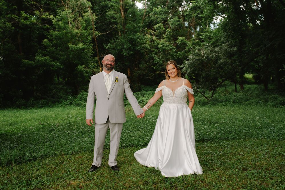 Bride and groom holding hands smiling in a lush green outdoor setting, bride in white gown, groom in tan suit.