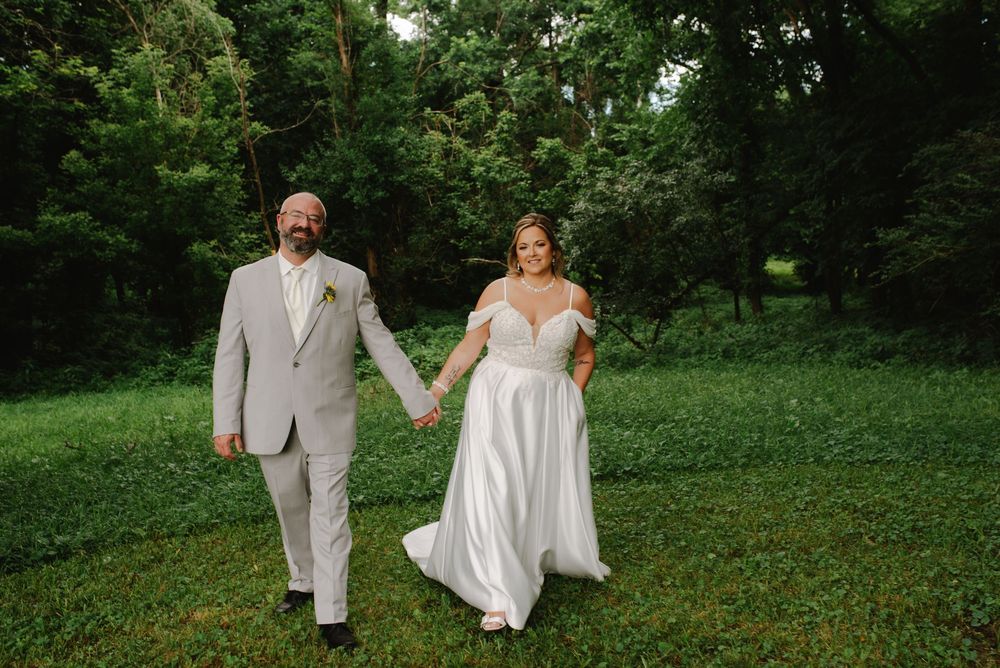 Bride in white gown and groom in gray suit hold hands walking through lush green garden on their wedding day.