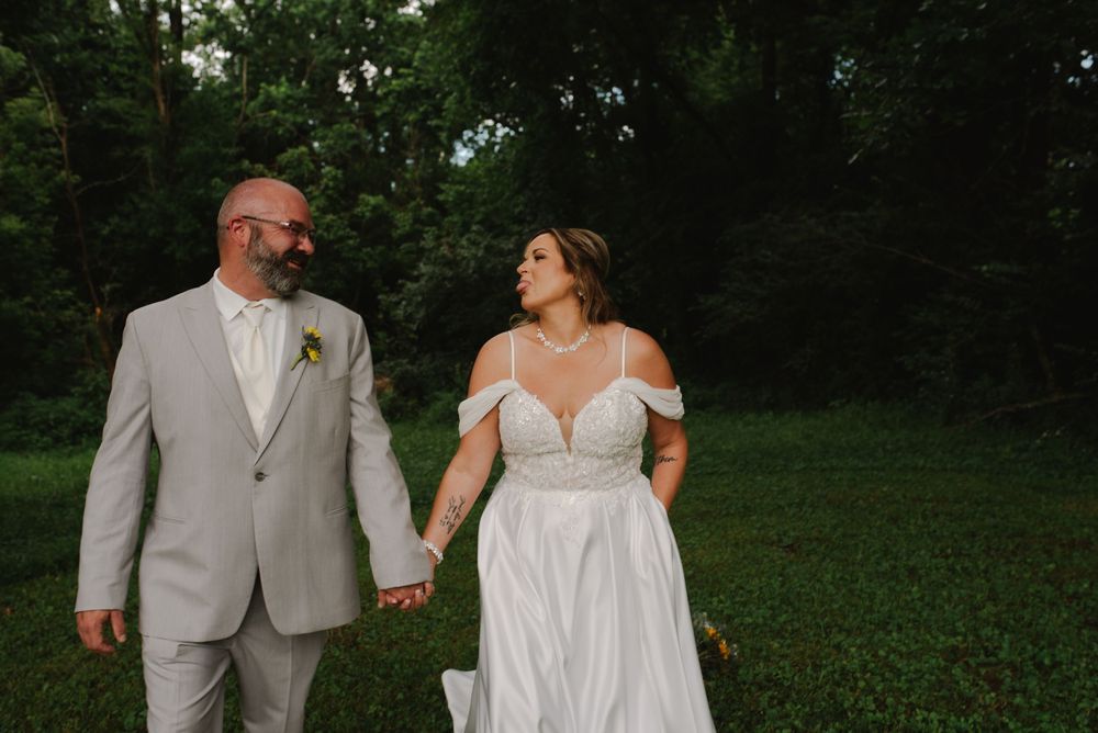 A bride in a white off-shoulder gown holds hands with a groom in a gray suit while walking through a lush green garden.
