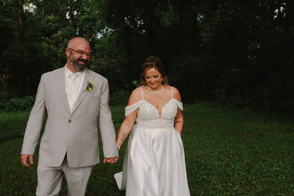 A bride and groom hold hands and smile while walking through a lush green outdoor setting on their wedding day.