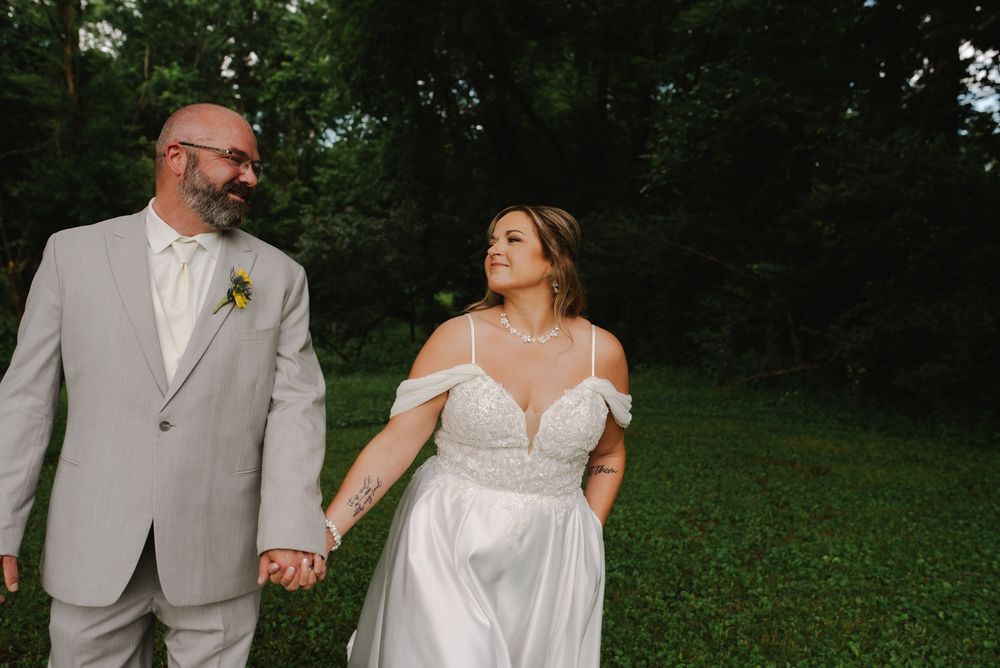 Bride in white off-shoulder gown and groom in gray suit hold hands walking through lush green outdoor wedding venue.