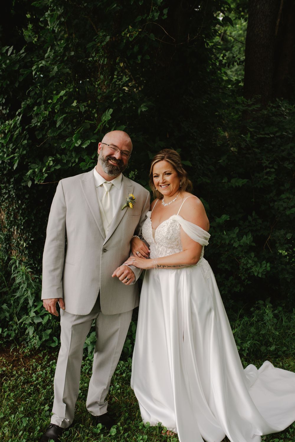 Smiling bride and groom pose together outdoors in lush greenery, wearing white gown and light gray suit with boutonniere.