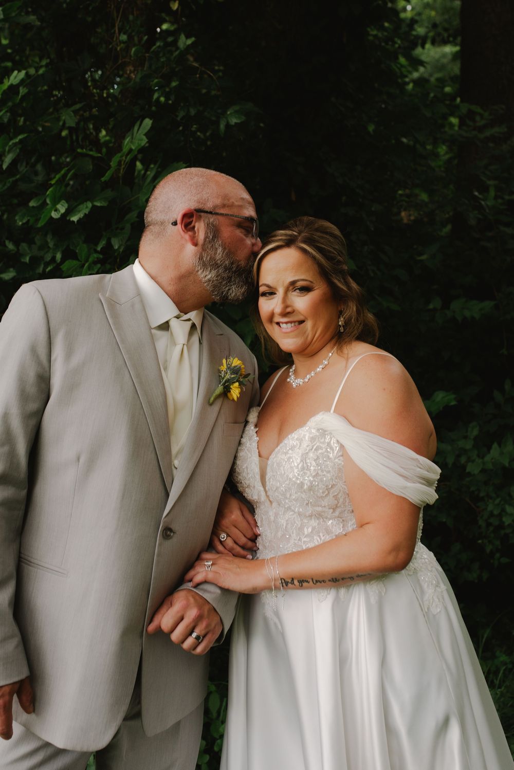 A bride in a white off-shoulder gown smiles as her groom in a tan suit whispers in her ear outdoors.