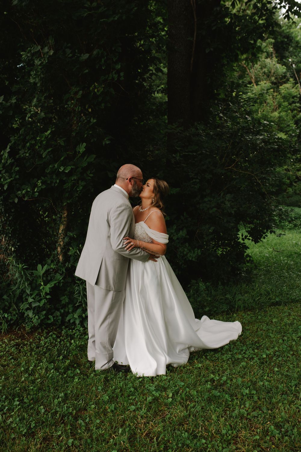 Bride and groom sharing a romantic kiss in a lush green forest setting, bride wearing an off-shoulder white gown.