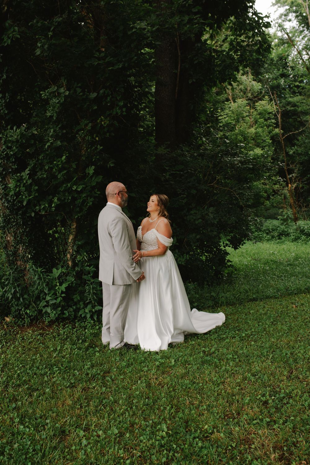 Bride and groom hold hands facing each other in a romantic outdoor garden setting surrounded by green trees.