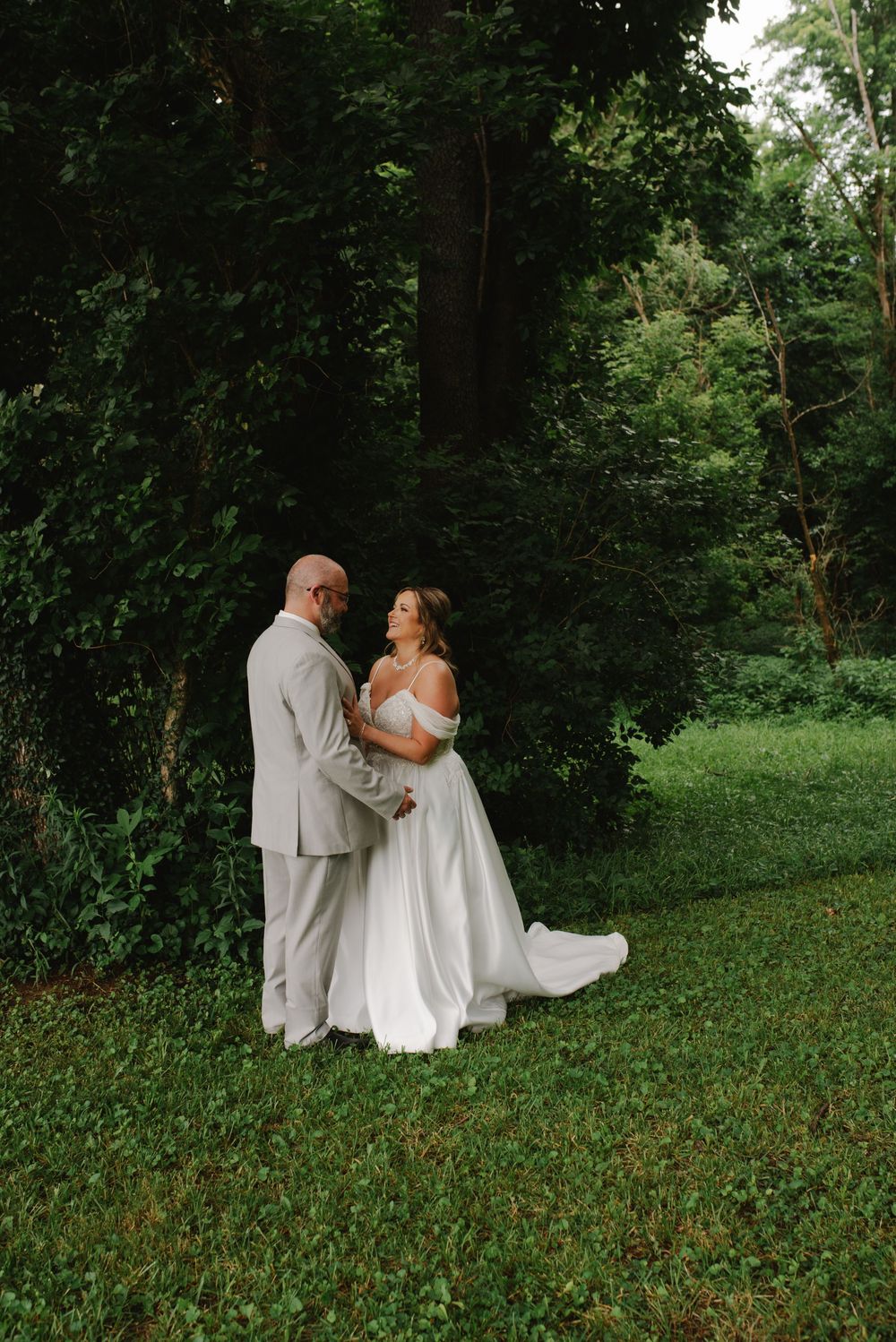 Bride and groom holding hands and gazing at each other in a green woodland garden, bride in elegant white dress.