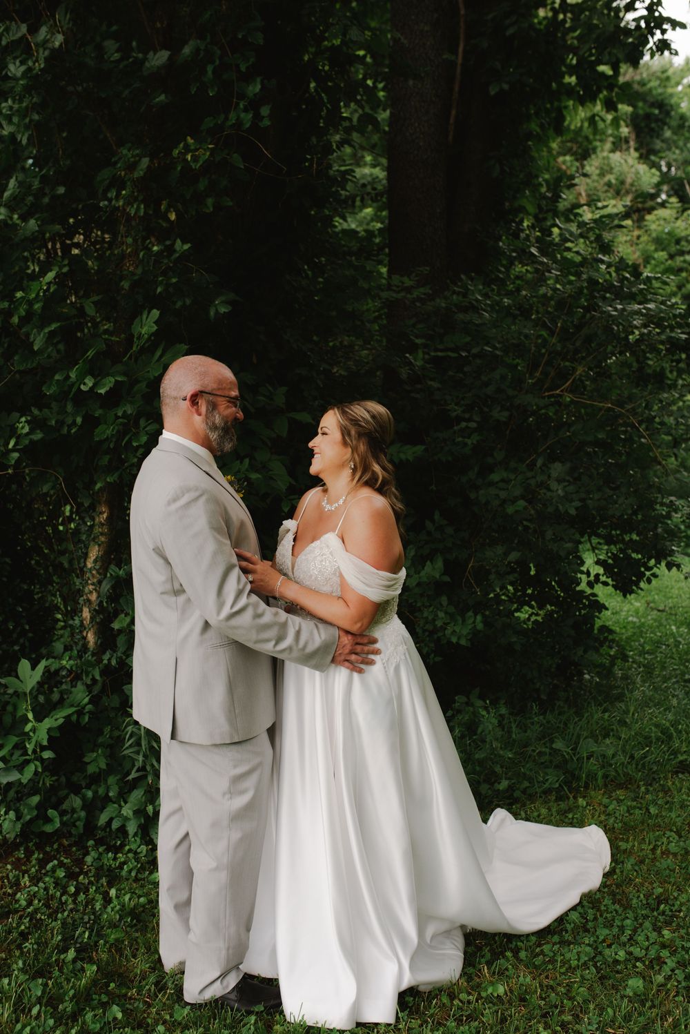 Bride and groom share a romantic embrace in front of lush green trees at an outdoor wedding.