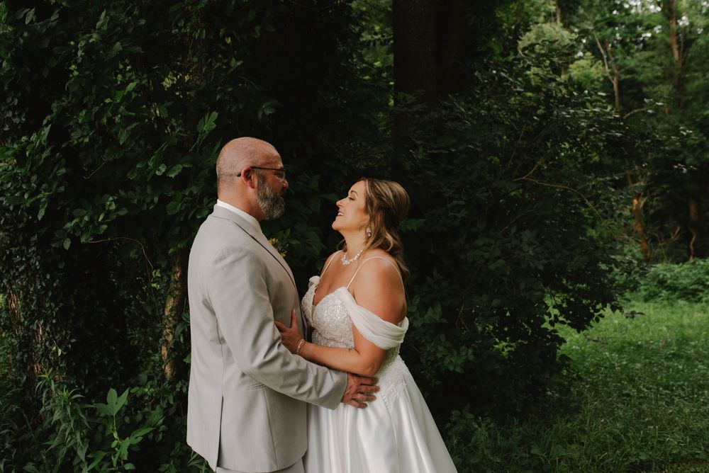 Bride and groom embrace in lush green garden setting, groom in gray suit, bride in off-shoulder gown.