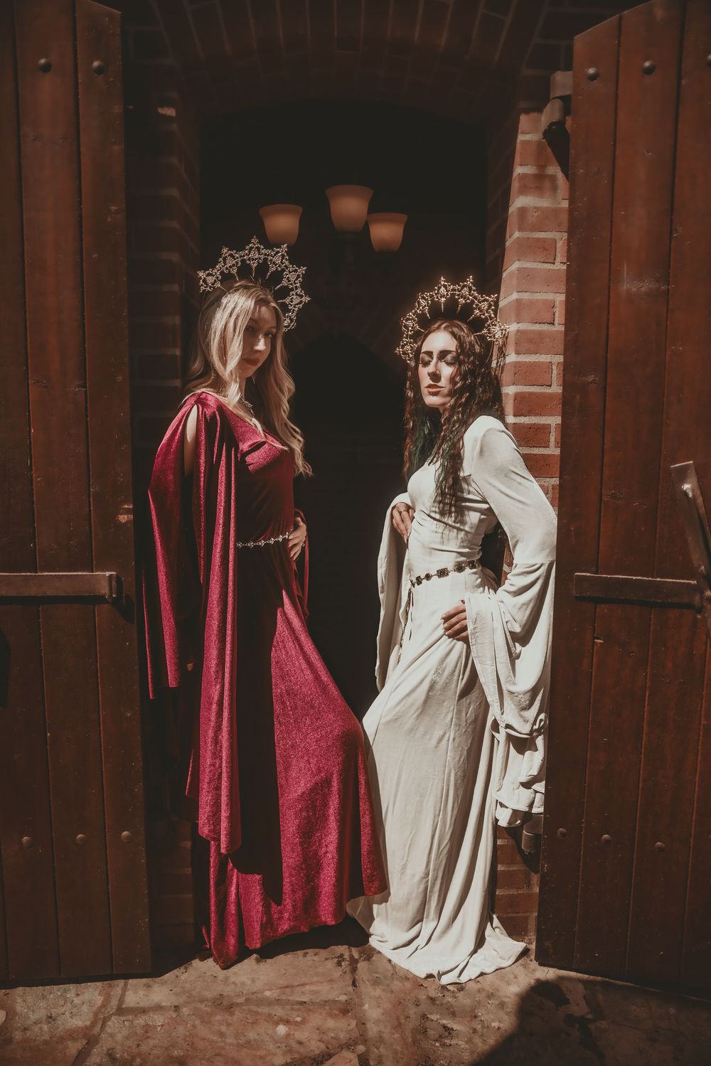 Two women in medieval gowns and crowns pose in a doorway, one in red velvet and one in white.