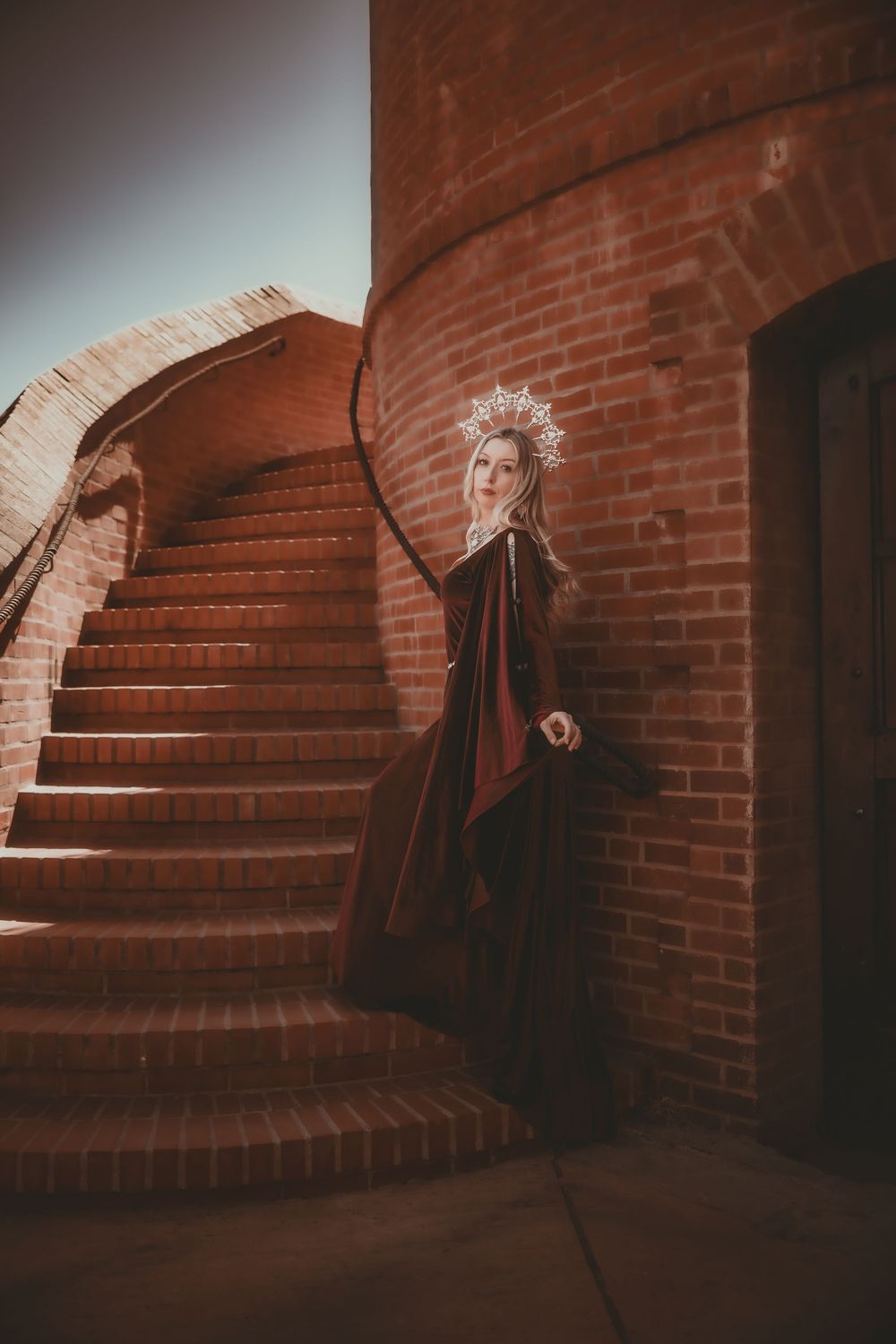 Woman in dark medieval gown and crown poses by brick spiral staircase in dramatic fantasy portrait.