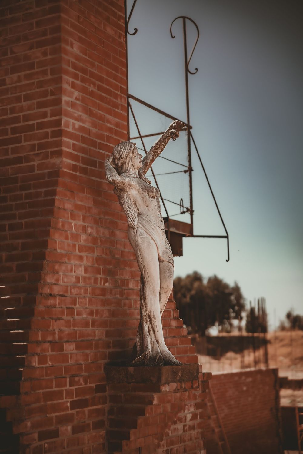 Weathered stone statue of a figure reaching upward beside a rustic red brick wall with metal brackets.