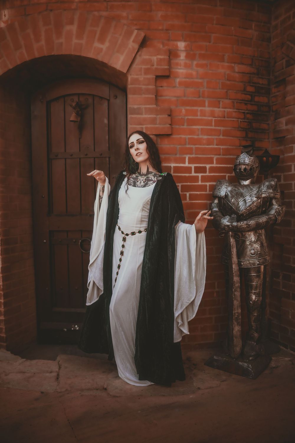 Woman in white medieval gown and black velvet cape poses beside suit of armor against red brick castle wall.