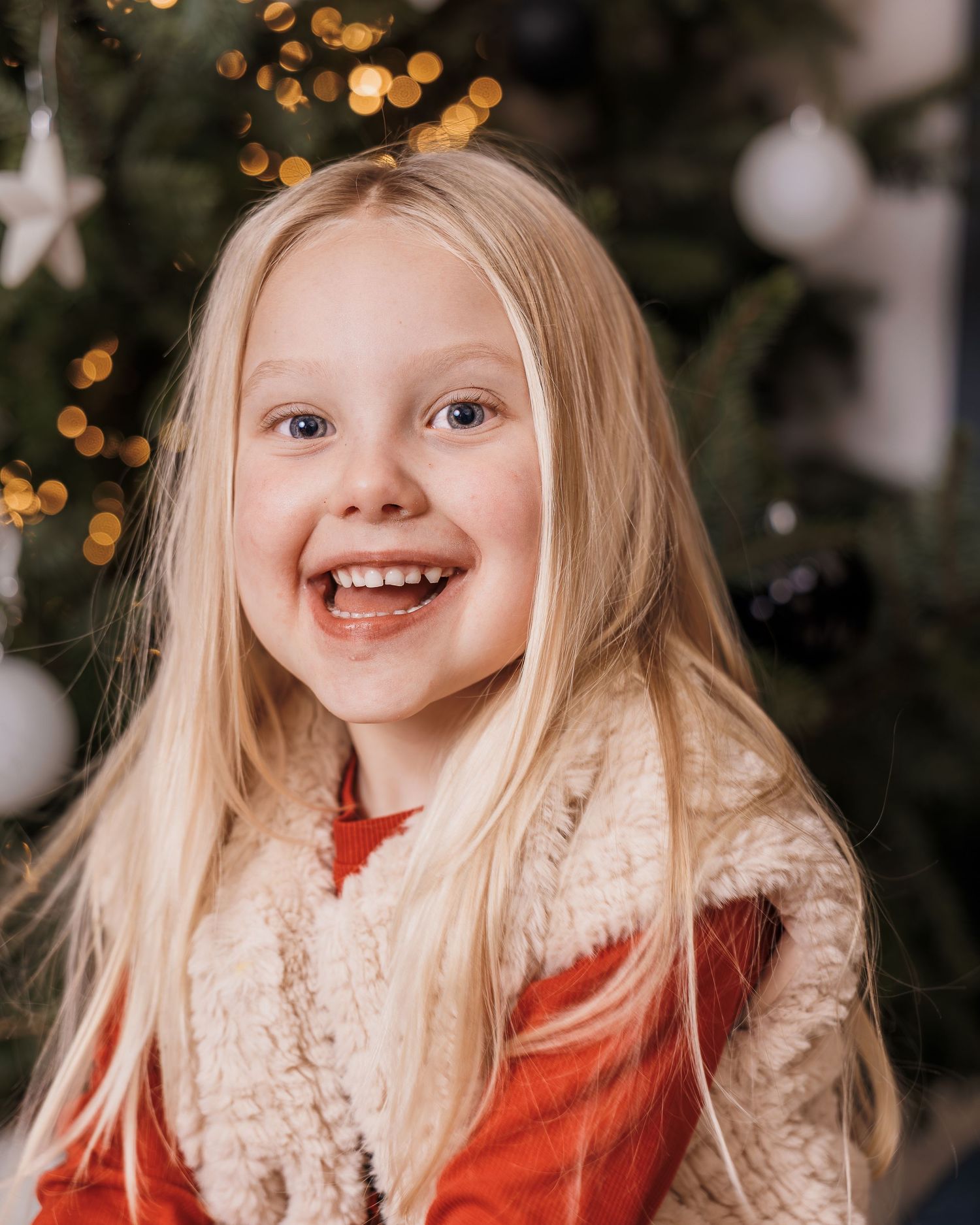 Smiling blonde girl in red outfit poses in front of a decorated Christmas tree with bokeh lights.
