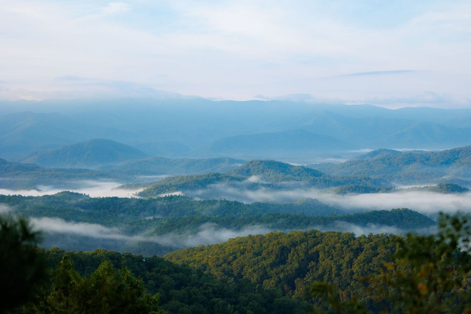 Aerial view of misty Blue Ridge Mountains with layers of rolling hills covered in lush green forest under a blue sky.