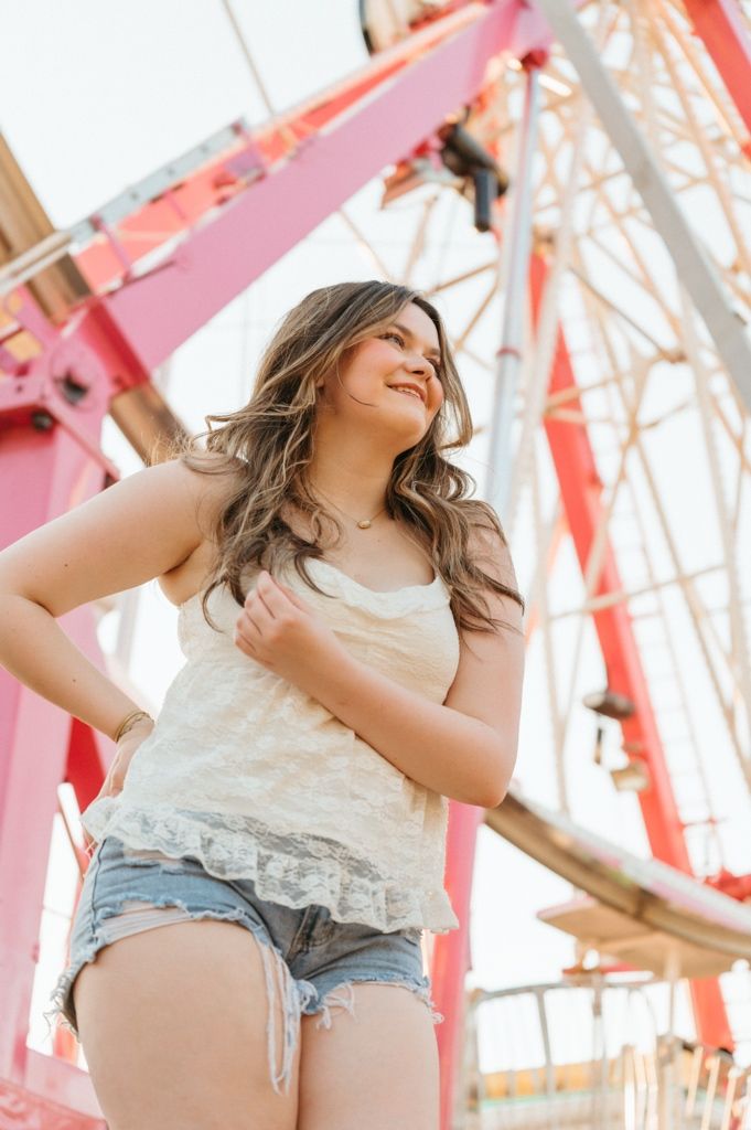 Young woman in denim shorts and lace tank top smiling near a pink Ferris wheel at an amusement park.