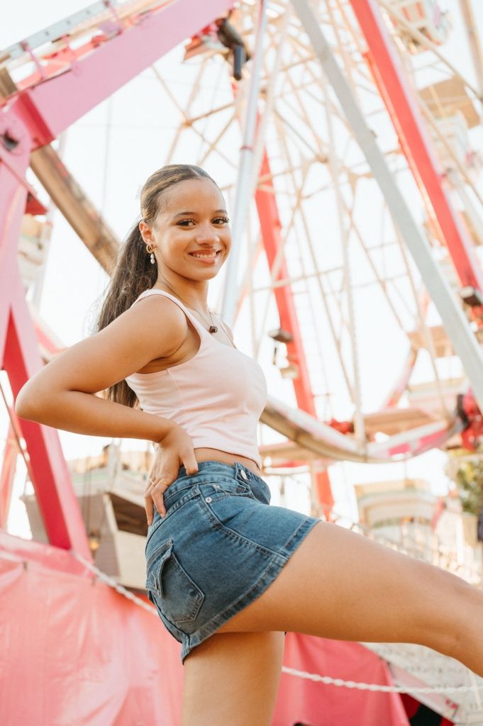 Smiling young woman in denim shorts poses at an amusement park with a pink Ferris wheel in the background.