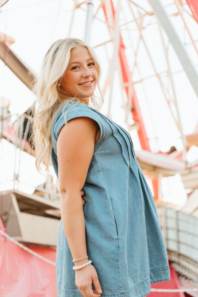 Smiling blonde woman in denim dress posing in front of a colorful Ferris wheel at an amusement park.