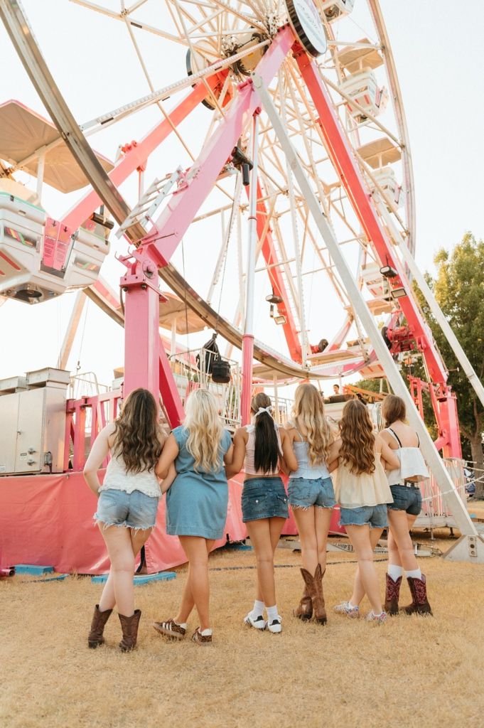 Six girls in denim shorts and cowboy boots stand before a pink Ferris wheel at a sunny county fair.