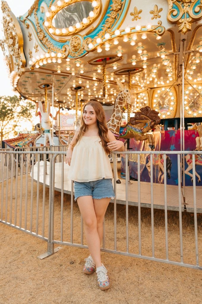 Young woman in white top and denim shorts smiling in front of a colorful lit carousel at a fairground.