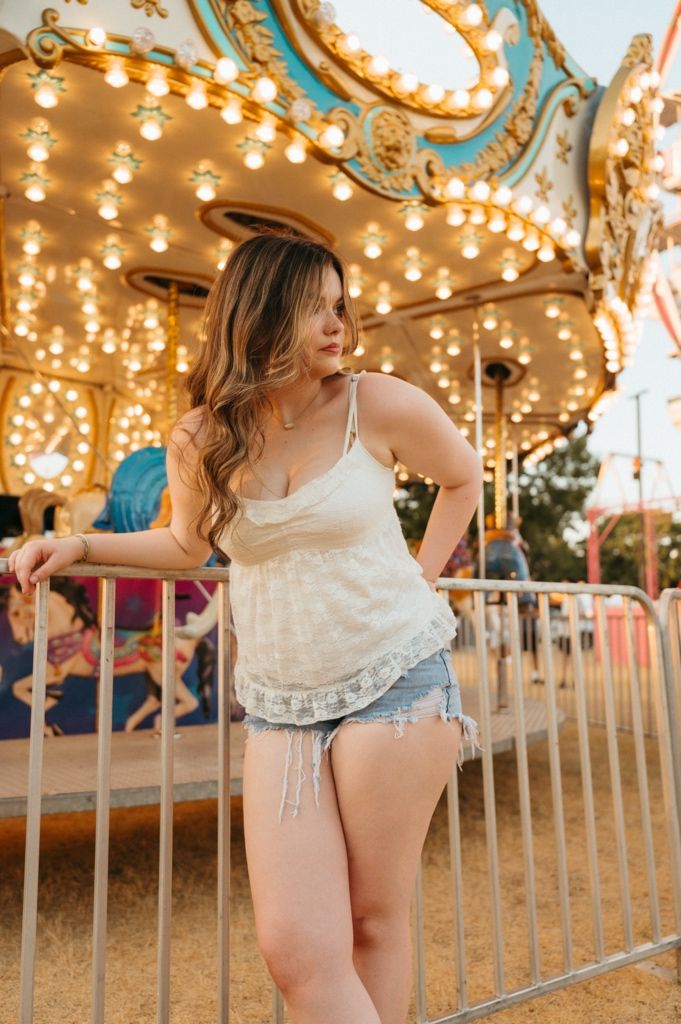 Young woman in white camisole and denim shorts posing by a lit carousel at a fairground during golden hour.