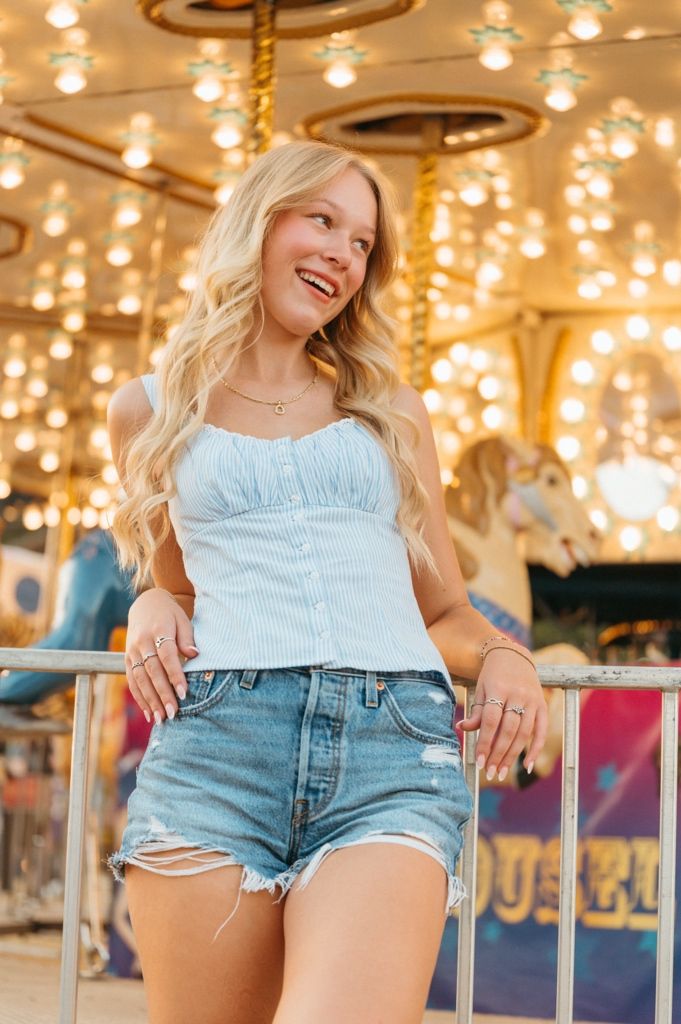 Smiling blonde woman in white top and denim shorts posing in front of a lit carousel at an amusement park.