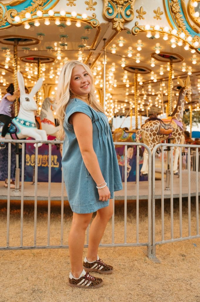 Smiling blonde woman in blue denim dress posing in front of illuminated carousel at amusement park.