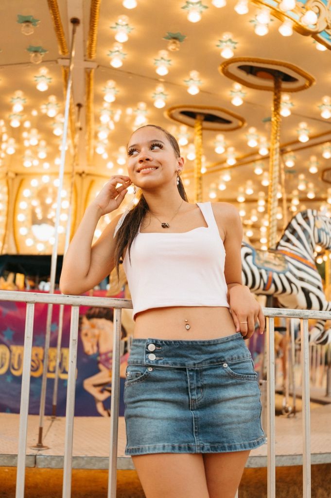 Young woman in white crop top and denim skirt smiling at a colorful carousel with bright lights at an amusement park.
