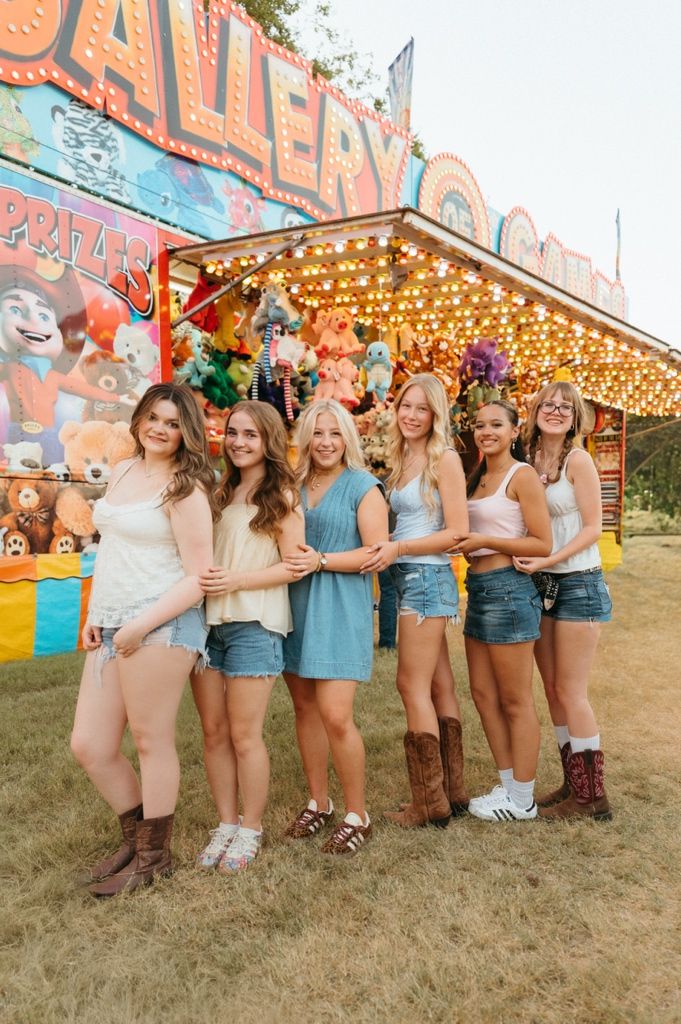 Six smiling girls in denim shorts pose together at a colorful carnival game booth with prize stuffed animals.
