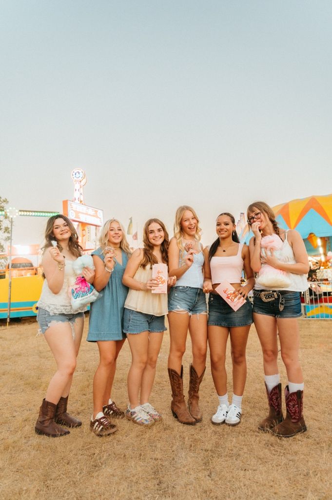 Six young women pose together at a county fair, wearing denim shorts and cowboy boots while holding fair treats.