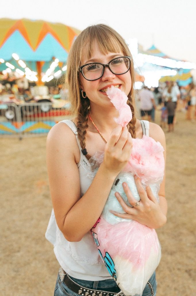 Young woman with braids and glasses eating pink cotton candy at a colorful outdoor carnival fair.