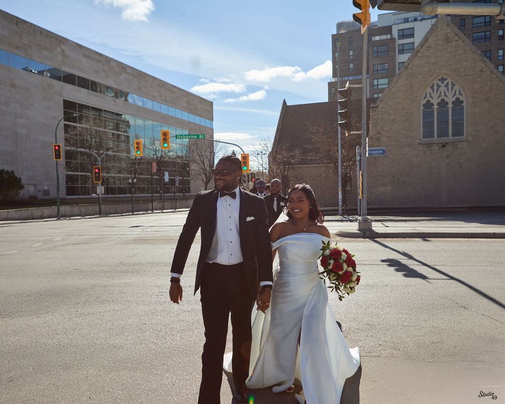 Newlywed couple in wedding attire walking across urban street intersection on sunny day.