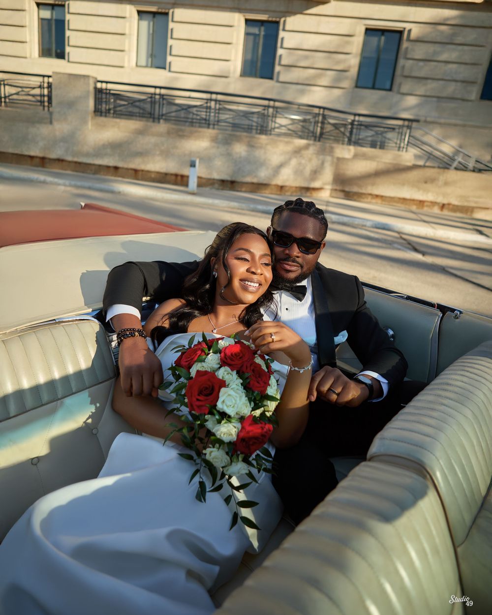 Joyful newlywed couple in wedding attire relaxing in a vintage convertible car, bride holding red roses bouquet.