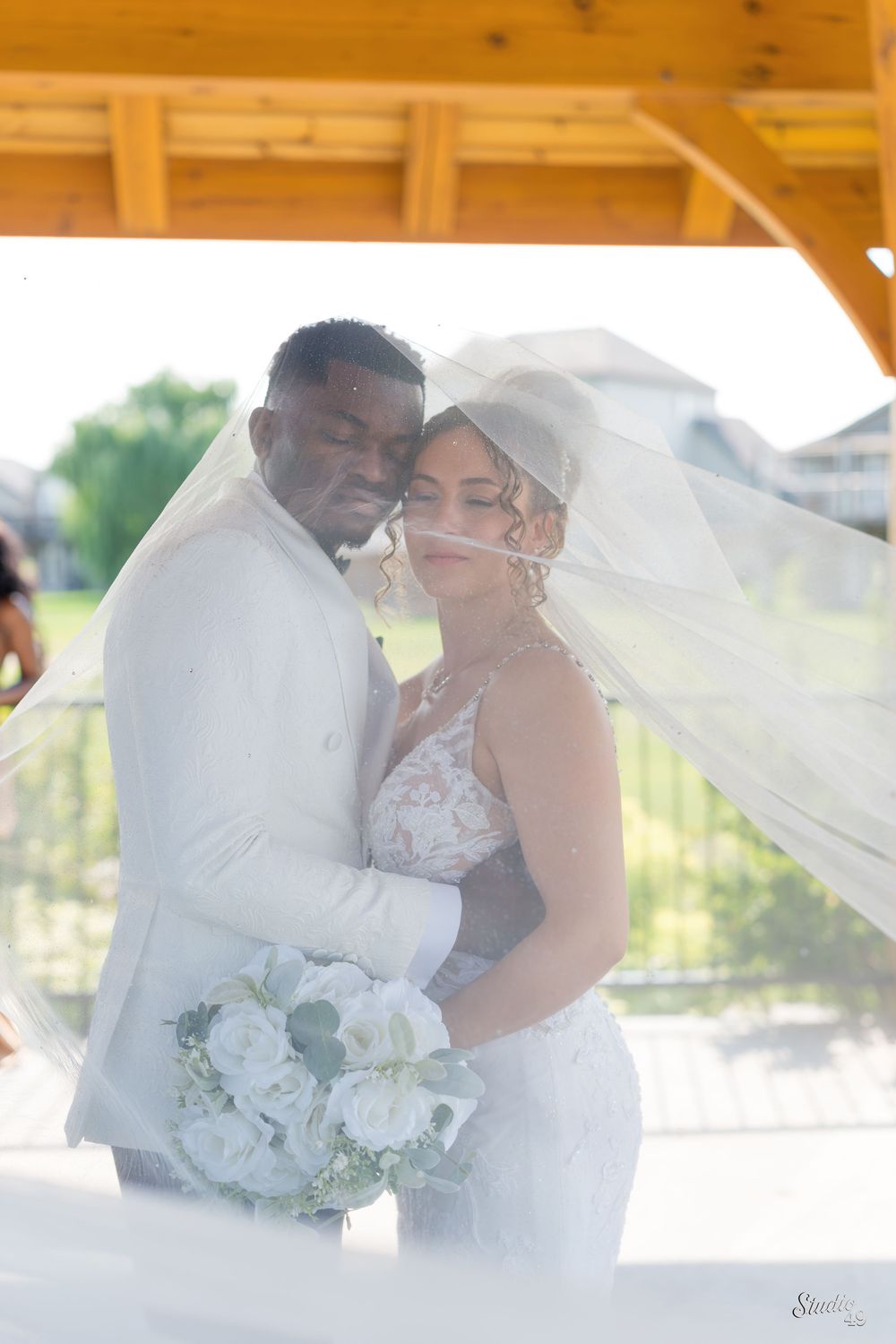 Bride and groom embracing under flowing veil with white bouquet at outdoor venue.