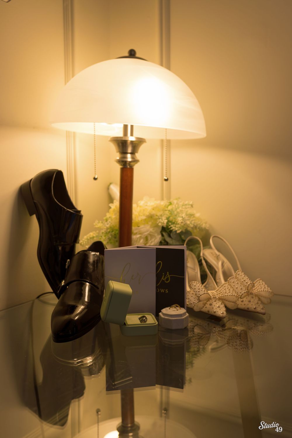 Wedding flat lay featuring black dress shoes, bridal heels, wedding rings, and vow books under a warm lamp.