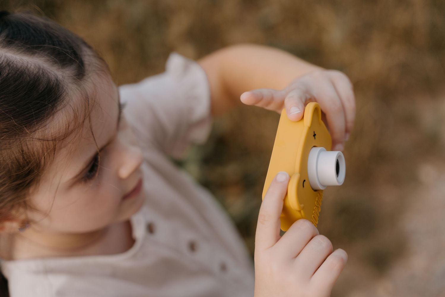 A young girl holds a small yellow toy camera, pointing at the lens with her finger outdoors.
