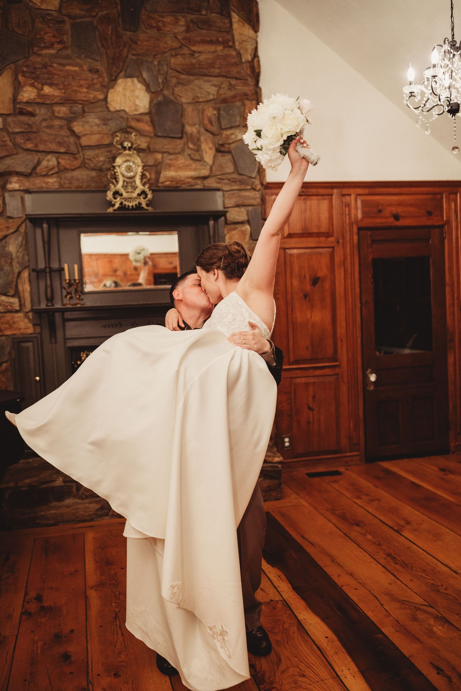 Groom lifting bride in white gown as she raises bouquet near stone fireplace in rustic wood-paneled room.
