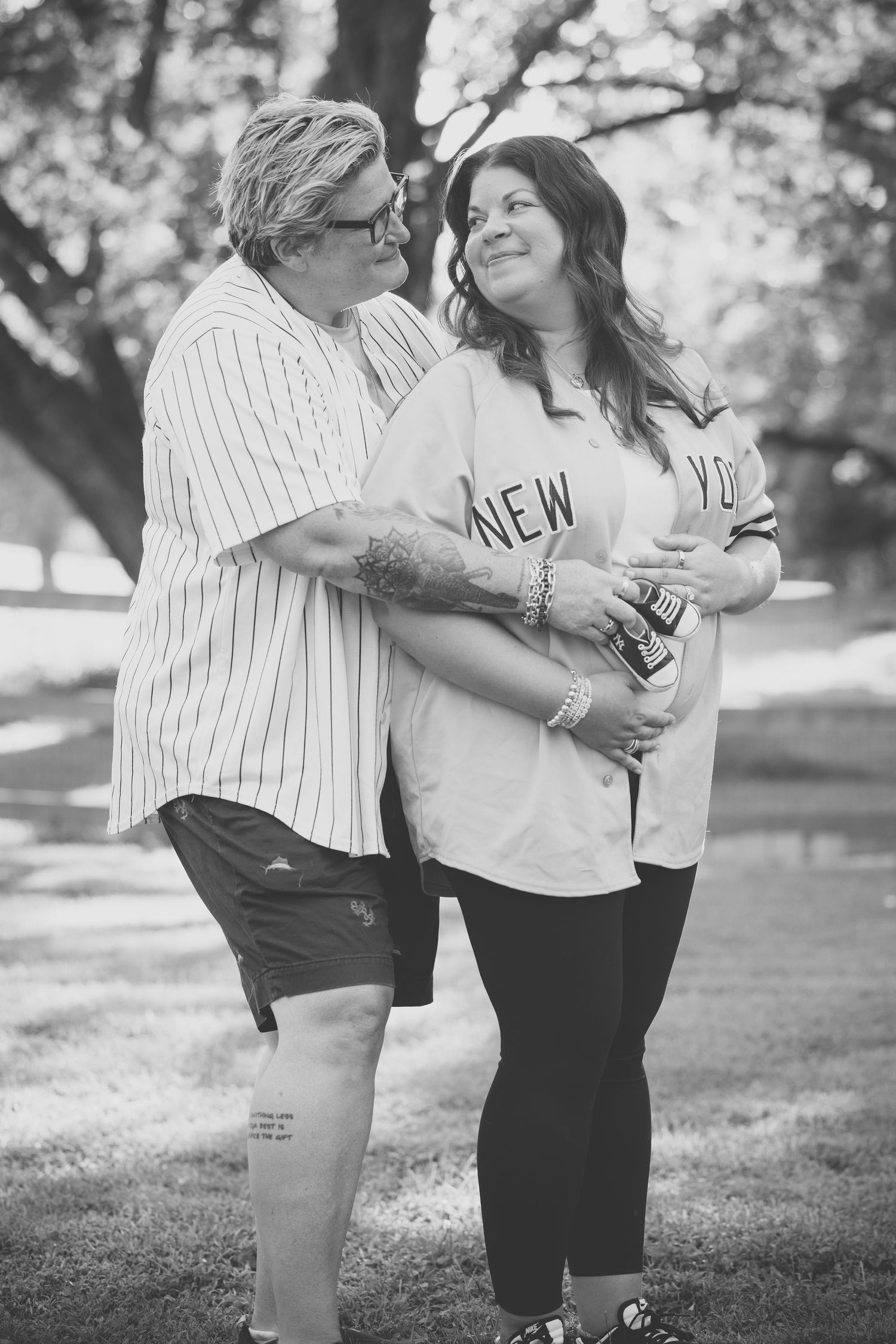 Two women holding ultrasound photos and a letter board pregnancy announcement sign outdoors in a park setting.
