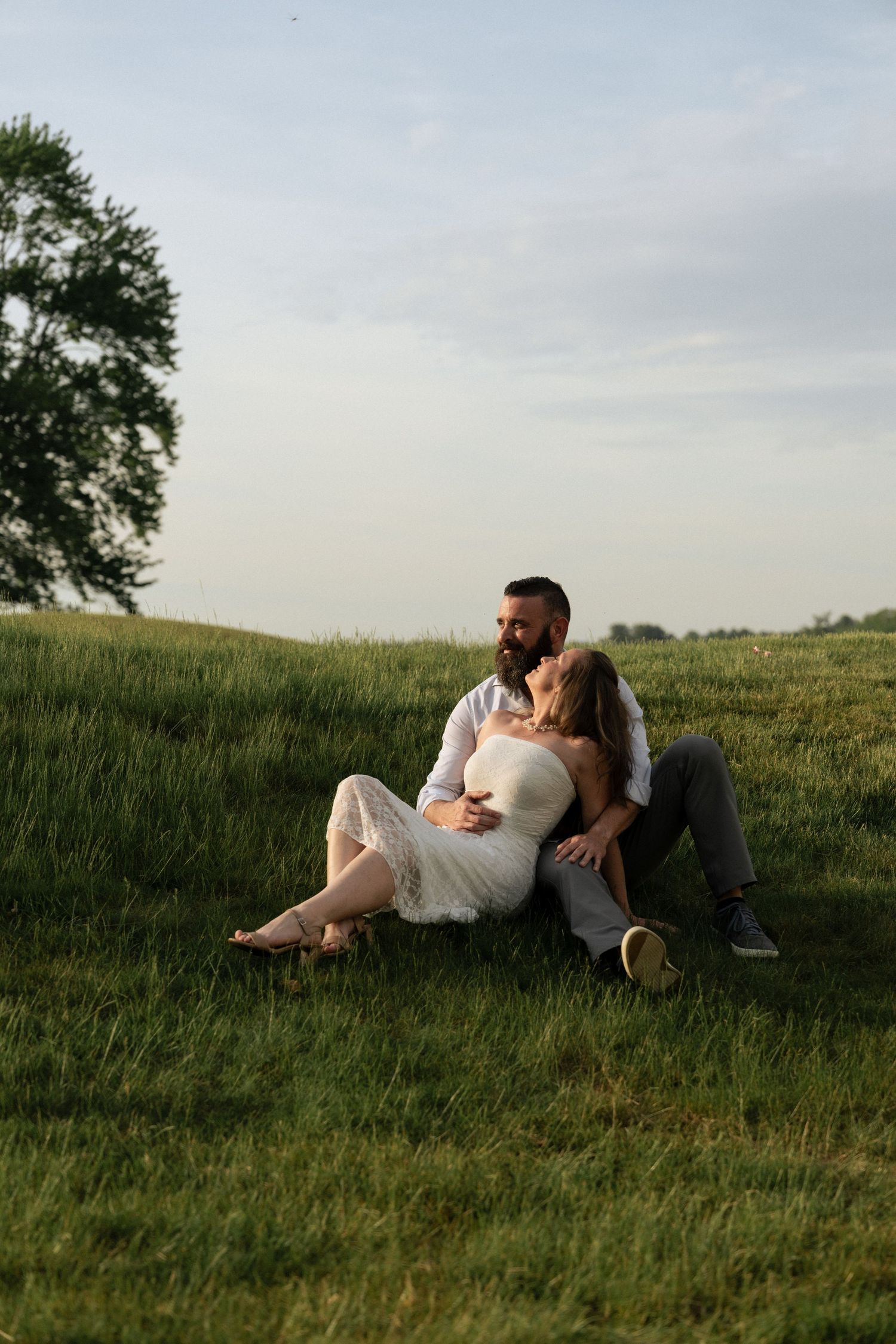 A couple sits together on a lush green hillside, the woman in a white dress leaning against her bearded partner at golden hour.