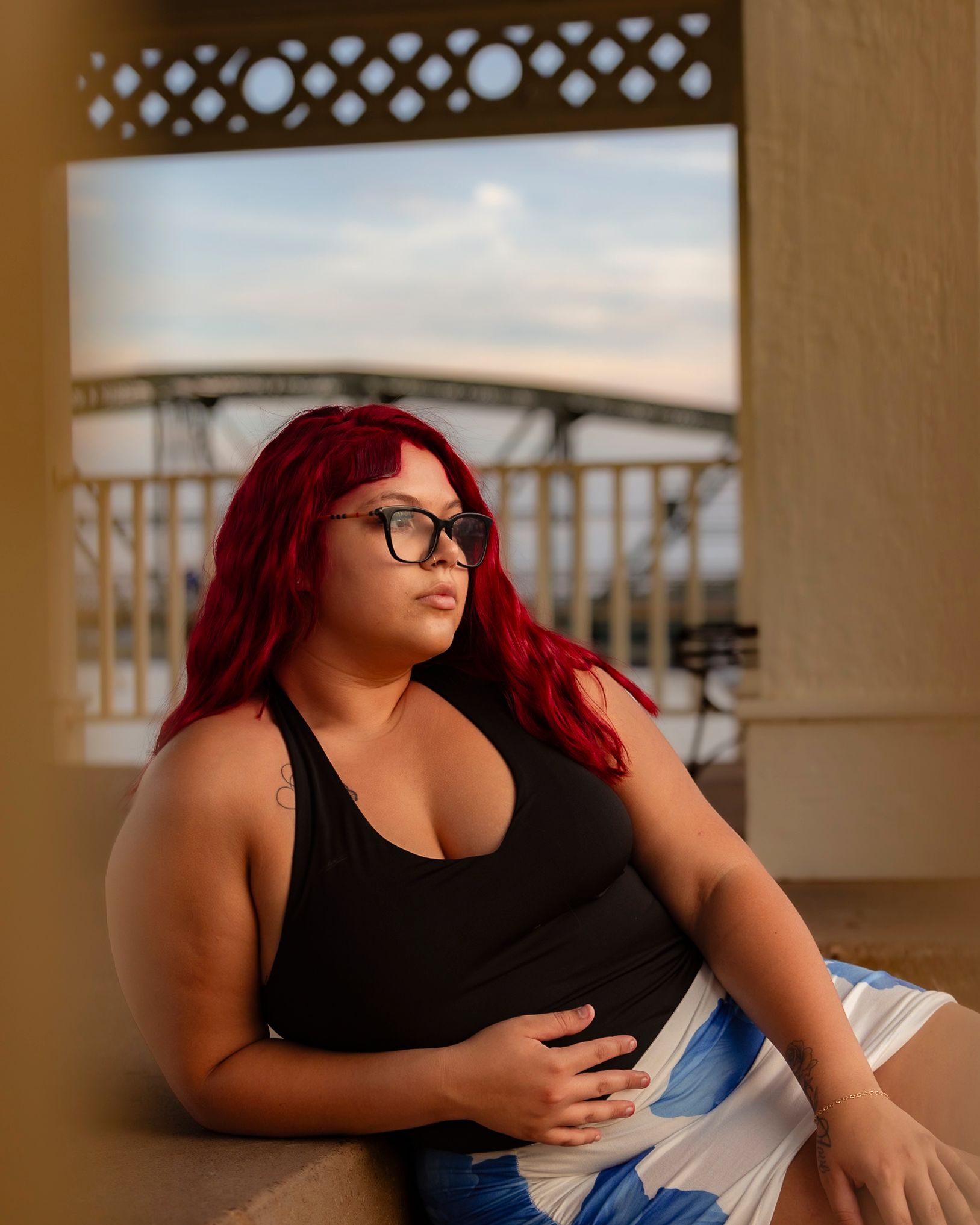 A young woman with vibrant red hair and glasses sits on a balcony, gazing thoughtfully into the distance.