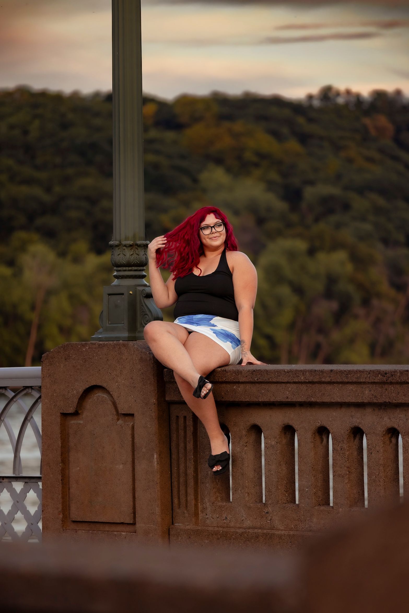 Red-haired woman in black top and denim skirt sitting on stone bridge railing with lush green trees in background.