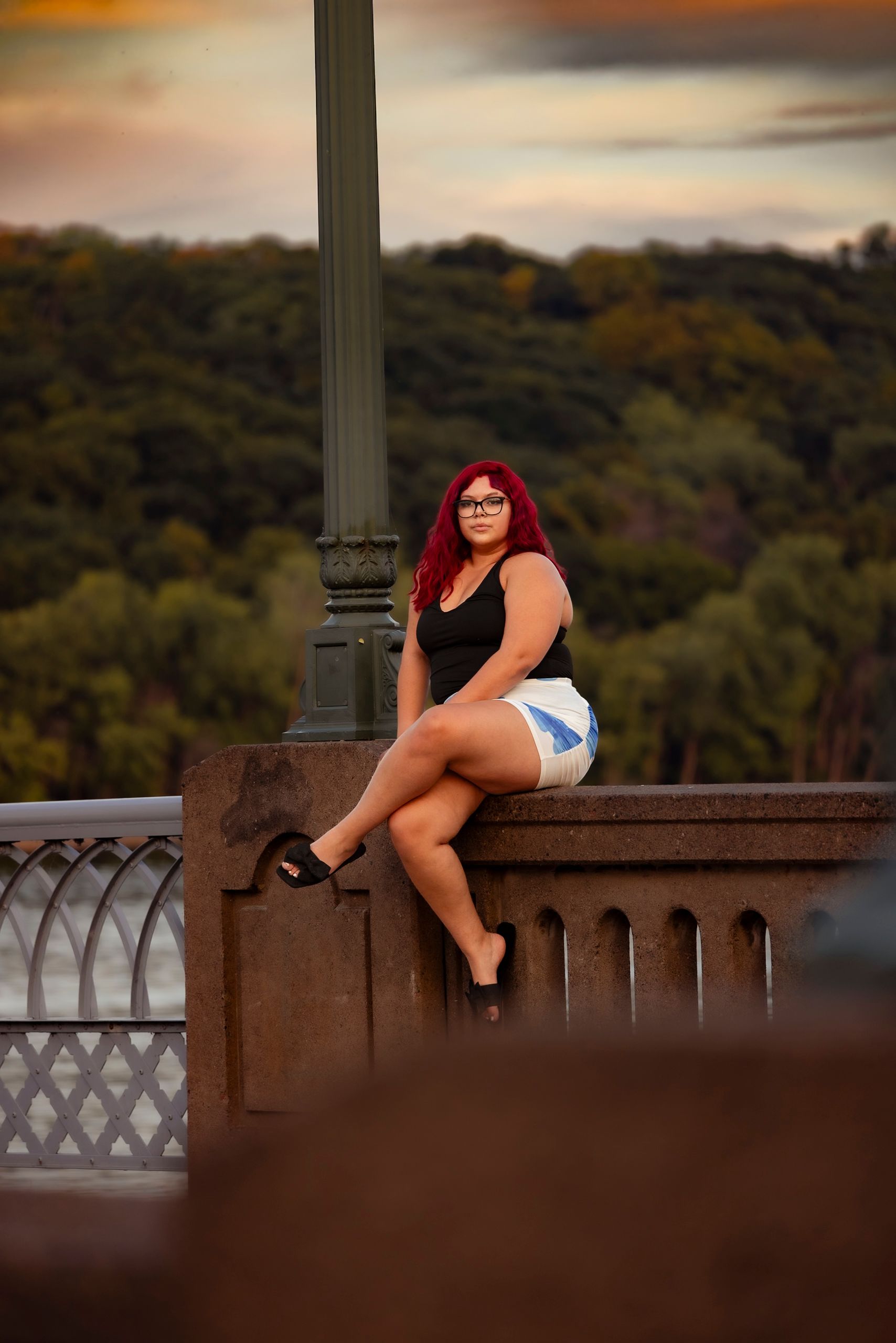 Red-haired woman in black top and denim shorts sitting on a stone bridge railing at sunset.