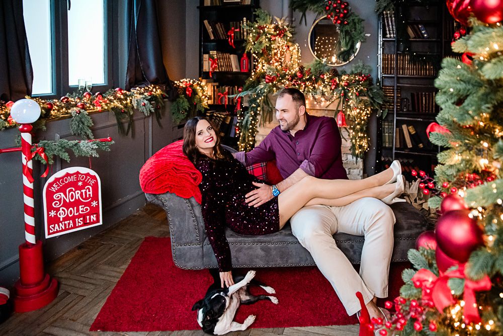 karácsonyi fotózás, páros
Couple sitting on a cozy sofa surrounded by festive Christmas decorations, lights, and a decorated tree indoors.