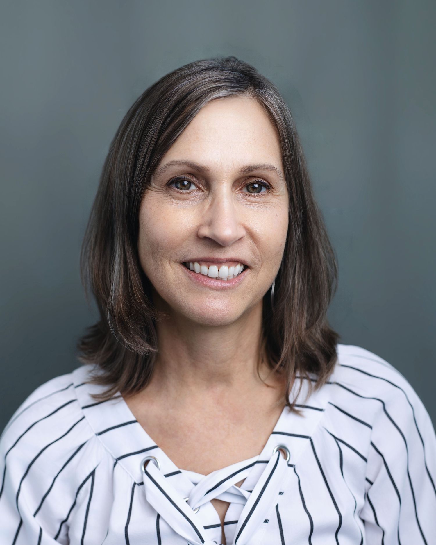 Professional headshot of a smiling brunette woman in a striped blouse on a gray background.