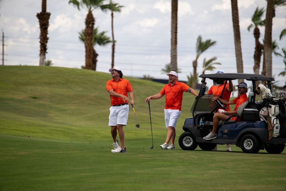 Two golfers in orange shirts stand on a lush golf course near a cart, with palm trees in the background.