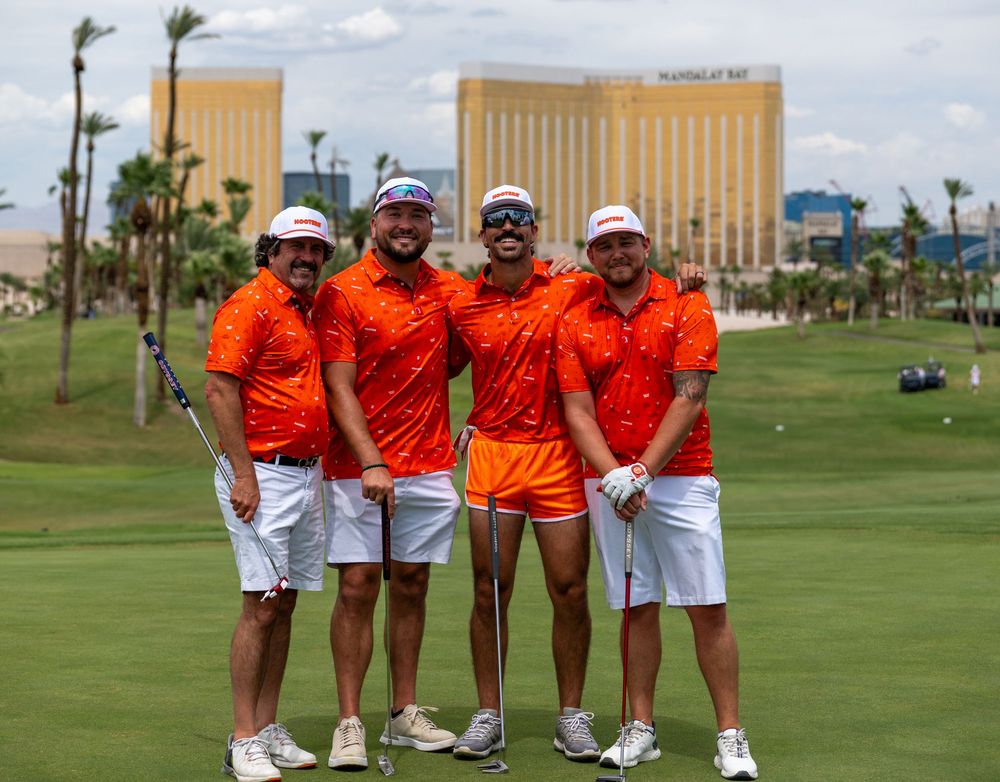 Four golfers in matching orange outfits pose on a Las Vegas golf course with the Mandalay Bay resort in the background.