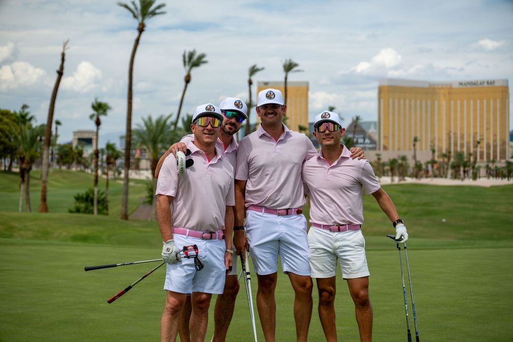 Four golfers in matching pink and white outfits pose together on a sunny golf course with a resort in the background.
