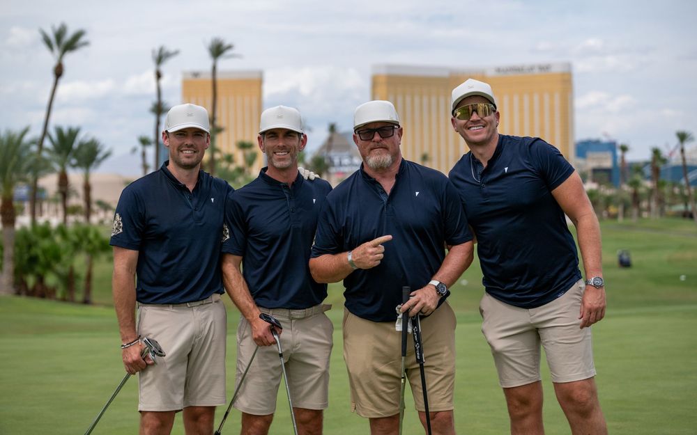 Four men in matching navy polo shirts and khaki shorts pose together on a golf course in Las Vegas.