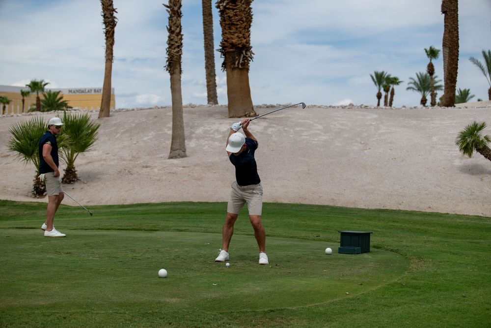Two golfers on a desert golf course, one mid-swing, with palm trees and sandy hills in the background.