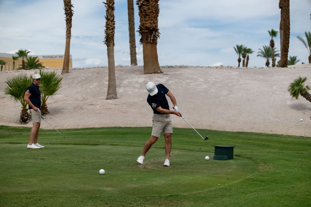 Two golfers practicing their swing on a lush green golf course with palm trees and sandy dunes in the background.